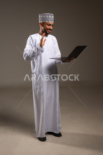الدردشة والتواصل مع الأهل والأصدقاء، إلقاء التحية والسلام، بورتريه لرجل عربي خليجي عماني يرتدي الدشداشة والكمة يقوم بإجراء مكالمة فيديو عبر الحاسوب المحمول يرفع يده بإيماءات الترحيب والسرور, استخدام التقنيات الحديثة المتطورة، الجسم بالطول الكامل، خلفية بيج