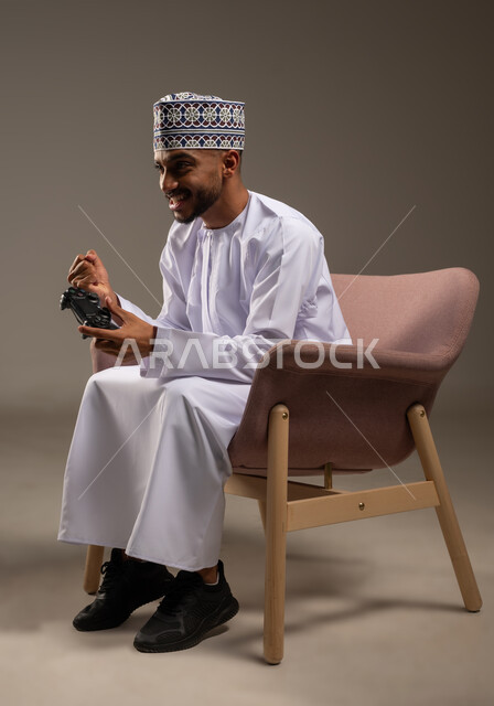 إيماءات الفوز والانتصار, بورتريه لرجل عربي خليجي عماني يرتدي الدشداشة والكمة يجلس على كرسي مريح يمسك عصا التحكم يرفع قبضة يده بتعابير الحماس، قضاء وقت ممتع في اللعب بألعاب الفيديو، مفهوم المتعة والتسلية، الاستمتاع بالتقنيات والتكنولوجيا الحديثة، خلفية بيج