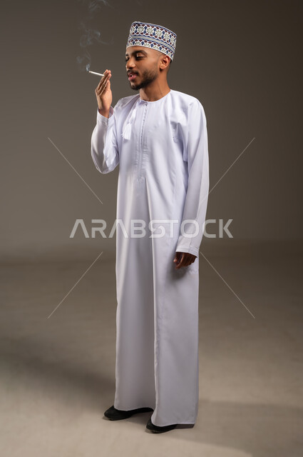 القيام بعادة سلوكية ضارة وتعاطي مادة سامة، الأثر السلبي للإدمان على التبغ والمخدرات، المعاناة من الاضطرابات النفسية والمشاكل الصحية، بورتريه لرجل عربي خليجي عماني يرتدي الدشداشة والكمة يقوم بتدخين السيجارة، الجسم بالطول الكامل، خلفية بيج