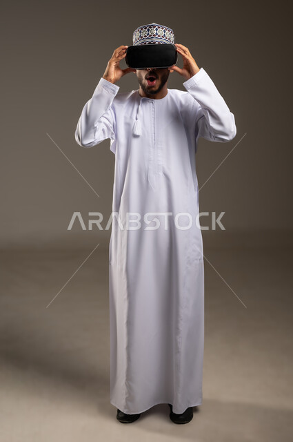 Opening mouth with gestures of shock and astonishment, portrait of an Arab Gulf Omani man wearing a dishdasha, a kimono and 3D glasses, merging and simulating the imaginary world, watching horror movies using virtual reality techniques, full-length body, white background