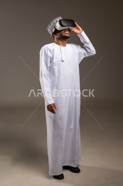 Integration and simulation of the imaginary world, fun and entertainment by watching through virtual reality glasses, portrait of a young Arab Gulf Omani man wearing a dishdasha, a kimono and 3D glasses (VR), technical and technological development, full-length body, white background