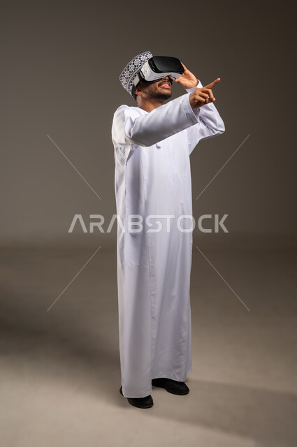 رفع السبابة للنقر على عناصر هولوجرامية، محاكاة العالم الخيالي، المتعة والتسلية بالمشاهدة عبر نظارة الواقع الافتراضي، بورتريه لشاب عربي خليجي عماني يرتدي الدشداشة والكمة ونظارات ثلاثية الأبعاد (الفي ار), التطور التقني والتكنولوجي, الجسم بالطول الكامل، خلفية بيضاء