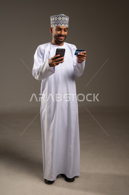 الدفع الإلكتروني عبر التقنيات الحديثة المتطورة، استخدام تطبيقات الجوال للدفع والشراء، بورتريه لشاب عربي خليجي عماني يرتدي الدشداشة والكمة يمسك بيده البطاقة الإلكترونية والهاتف المحمول ينظر بإيماءات البشاشة والسرور, الجسم بالطول الكامل، خلفية بيج