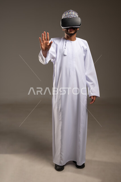 Technical and technological development, fun and entertainment watching through virtual reality glasses, portrait of a young Arab Gulf Omani man wearing a dishdasha, a kimono and 3D glasses (VR) raising his hand to touch holographic elements, integration and simulation of the imaginary world, full-length body, white background