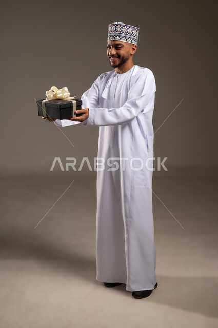 Giving gifts on Eid and special occasion, expressing love, appreciation and respect, portrait of an Arab Gulf Omani man wearing a dishdasha and a kummah holding a gift box looking at something with gestures of pleasure and happiness, concept of thanks and gratitude, full length body, beige background