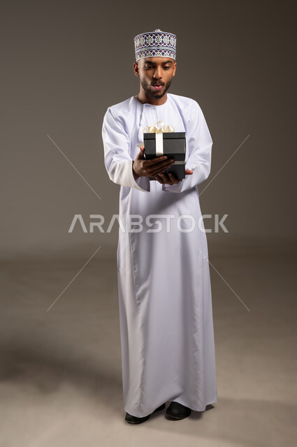 إيماءات الذهول والمفاجأة، تقديم الهدايا في العيد والمناسبة الخاصة، التعبير عن الحب والتقدير والاحترام، بورتريه لرجل عربي خليجي عماني يرتدي الدشداشة والكمة يحمل صندوق الهدية ينظر إليه بتعابير السرور والحماس، مفهوم الشكر والامتنان، الجسم بالطول الكامل، خلفية بيج
