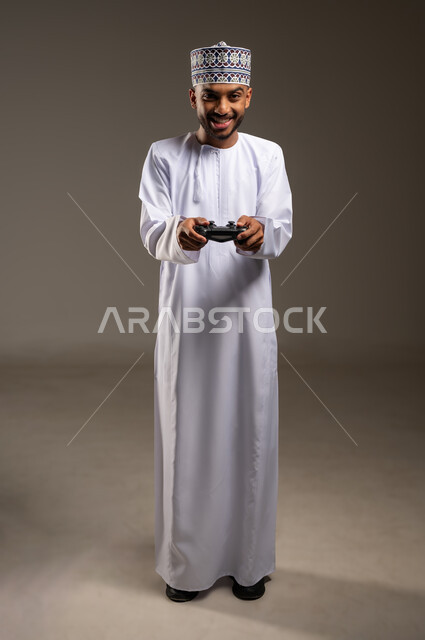 الاستمتاع بالتقنيات والتكنولوجيا الحديثة، إيماءات التفاعل والاندماج, بورتريه لرجل عربي خليجي عماني يرتدي الدشداشة والكمة يمسك عصا التحكم ينظر إلى الكاميرا بتعابير السرور، مفهوم المتعة والتسلية، قضاء وقت ممتع في اللعب بألعاب الفيديو، الجسم بالطول الكامل، خلفية بيج