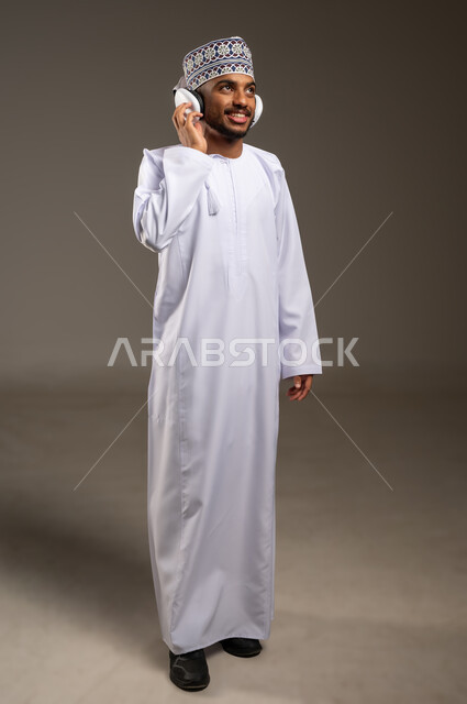 التفاعل مع الأنغام الموسيقية باستخدام الهيدفون، إيماءات الاندماج مع الأغنية، بورتريه لرجل عربي خليجي عماني يرتدي الدشداشة والكمة يستمع إلى الموسيقى من خلال سماعة الرأس بتعابير الانصات والاستمتاع، موهبة غنائية فنية، الجسم بالطول الكامل، خلفية بيج