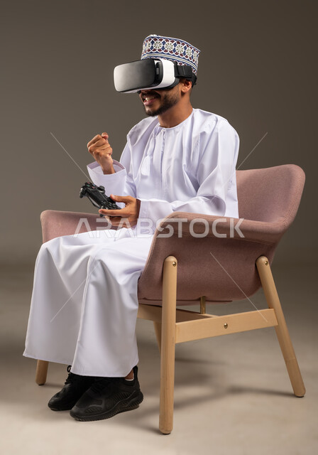 ايماءات الفوز والنجاح، محاكاة العالم الخيالي، التفاعل والاندماج مع ألعاب الفيديو، قضاء وقت ممتع ومسلي، بورتريه لرجل عربي خليجي عماني يرتدي الدشداشة والكمة ونظارة الواقع الافتراضي يجلس على كرسي مريح يحمل عصا التحكم يرفع يده بتعابير النصر، خلفية بيضاء