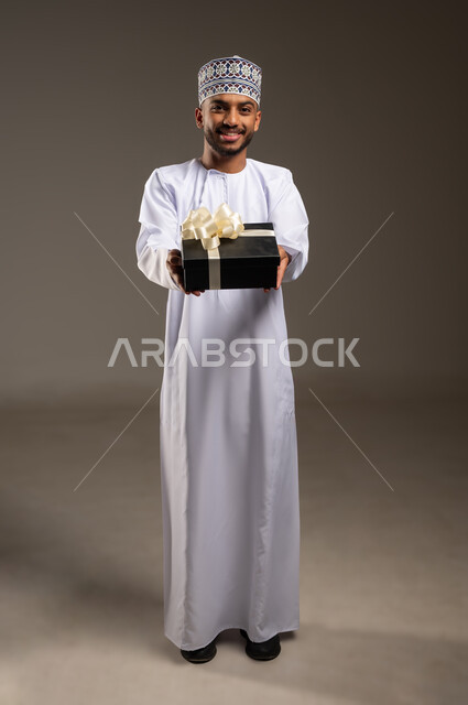 التعبير عن الحب والتقدير والاحترام، بورتريه لرجل عربي خليجي عماني يرتدي الدشداشة والكمة يحمل صندوق الهدية ينظر إلى الكاميرا بإيماءات السرور، تقديم الهدايا في العيد والمناسبة الخاصة، مفهوم الشكر والامتنان، الجسم بالطول الكامل، خلفية بيج