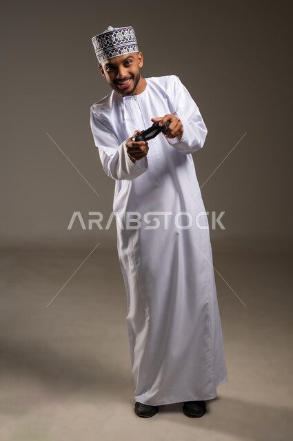إيماءات التفاعل والاندماج, بورتريه لرجل عربي خليجي عماني يرتدي الدشداشة والكمة يمسك عصا التحكم ينظر إلى الكاميرا بتعابير السرور، مفهوم المتعة والتسلية، قضاء وقت ممتع في اللعب بألعاب الفيديو، الاستمتاع بالتقنيات والتكنولوجيا الحديثة، الجسم بالطول الكامل، خلفية بيج