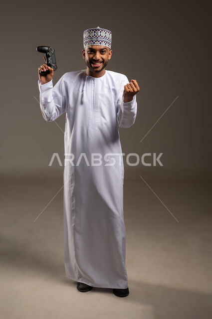 قضاء وقت ممتع في اللعب بألعاب الفيديو، إيماءات الفوز والانتصار, بورتريه لرجل عربي خليجي عماني يرتدي الدشداشة والكمة يمسك عصا التحكم يرفع قبضة يده بتعابير الحماس، مفهوم المتعة والتسلية، الاستمتاع بالتقنيات والتكنولوجيا الحديثة، الجسم بالطول الكامل، خلفية بيج