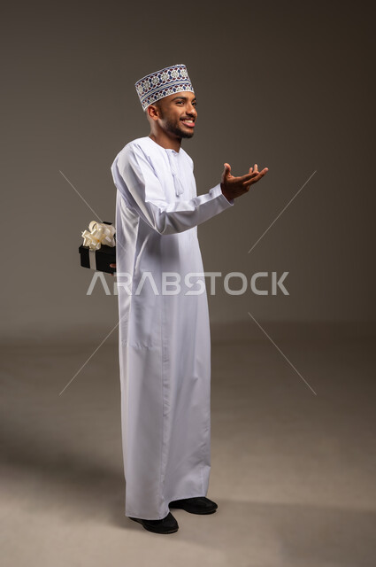 مفهوم الشكر والامتنان، التعبير عن الحب والتقدير والاحترام، بورتريه لرجل عربي خليجي عماني يرتدي الدشداشة والكمة يحمل صندوق الهدية خلف ظهره ينظر إلى شيء ما بإيماءات السرور، تقديم الهدايا في العيد والمناسبة الخاصة، الجسم بالطول الكامل، خلفية بيج