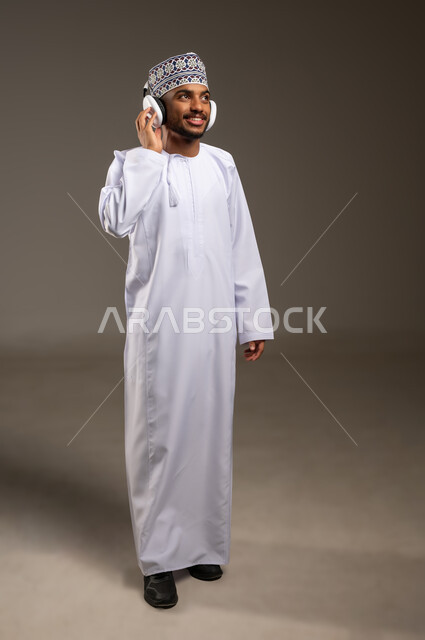 إيماءات الاندماج مع الأغنية، بورتريه لرجل عربي خليجي عماني يرتدي الدشداشة والكمة يستمع إلى الموسيقى من خلال سماعة الرأس بتعابير الانصات والاستمتاع، التفاعل مع الأنغام الموسيقية باستخدام الهيدفون، موهبة غنائية فنية، الجسم بالطول الكامل، خلفية بيج