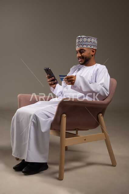 استخدام تطبيقات الجوال للدفع والشراء، بورتريه لشاب عربي خليجي عماني يرتدي الدشداشة والكمة يجلس على كرسي مريح يمسك بيده البطاقة الإلكترونية والهاتف المحمول, إمكانية الدفع الإلكتروني عبر التقنيات الحديثة المتطورة، خلفية بيج