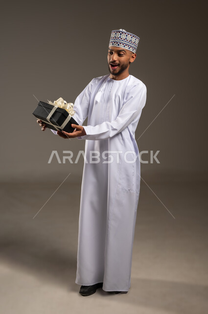 تقديم الهدايا في العيد والمناسبة الخاصة، مفهوم الشكر والامتنان، التعبير عن الحب والتقدير والاحترام، بورتريه لرجل عربي خليجي عماني يرتدي الدشداشة والكمة يحمل صندوق الهدية ينظر إليه بايماءات المفاجأة والصدمة، الجسم بالطول الكامل، خلفية بيج
