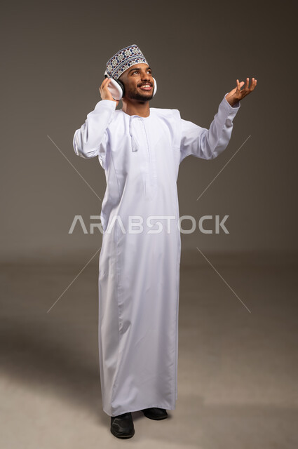 موهبة غنائية فنية، إيماءات الاندماج مع الأغنية، بورتريه لرجل عربي خليجي عماني يرتدي الدشداشة والكمة يستمع إلى الموسيقى من خلال سماعة الرأس يرفع يده بتعابير الانصات والاستمتاع، التفاعل مع الأنغام الموسيقية باستخدام الهيدفون، الجسم بالطول الكامل، خلفية بيج
