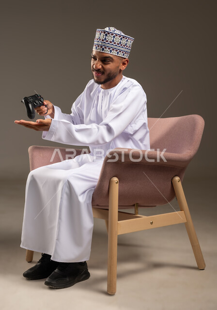 The concept of fun and entertainment, gestures of integration and interaction, portrait of an Arab Gulf Omani man wearing a dishdasha and a turban sitting on a comfortable chair holding a joystick in his hand, having a good time playing video games, enjoying modern techniques and technology, beige background
