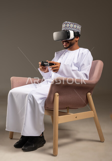 اللعب بألعاب الفيديو الترفيهية، استخدام الأجهزة التكنولوجية والتقنيات الحديثة، بورتريه لرجل عربي خليجي عماني يرتدي الدشداشة والكمة ونظارة الواقع الإفتراضي يجلس على كرسي مريح يمسك بيده عصا التحكم، دمج العالم الخيالي مع المتعة والتسلية، خلفية بيج