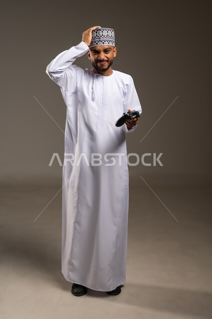 إيماءات الخسارة والخيبة, بورتريه لرجل عربي خليجي عماني يرتدي الدشداشة والكمة يمسك عصا التحكم يضع يده على رأسه بتعابير الحزن والاستياء، قضاء وقت ممتع في اللعب بألعاب الفيديو، الاستمتاع بالتقنيات والتكنولوجيا الحديثة، الجسم بالطول الكامل، خلفية بيج