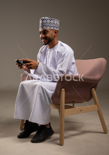 الاستمتاع بالتقنيات والتكنولوجيا الحديثة، مفهوم المتعة والتسلية، إيماءات الاندماج والتفاعل, بورتريه لرجل عربي خليجي عماني يرتدي الدشداشة والكمة يجلس على كرسي مريح يمسك بيده عصا التحكم، قضاء وقت ممتع في اللعب بألعاب الفيديو، خلفية بيج
