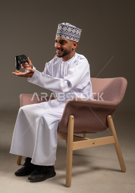 قضاء وقت ممتع في اللعب بألعاب الفيديو، الاستمتاع بالتقنيات والتكنولوجيا الحديثة، مفهوم المتعة والتسلية، إيماءات الاندماج والتفاعل, بورتريه لرجل عربي خليجي عماني يرتدي الدشداشة والكمة يجلس على كرسي مريح يمسك بيده عصا التحكم، خلفية بيج