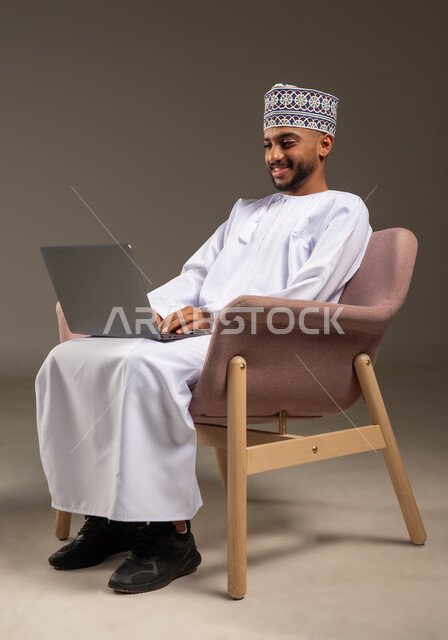 إدارة شؤون العمل وتنظيمه، بورتريه لرجل عربي خليجي عماني يرتدي الدشداشة والكمة يجلس على كرسي مريح يعمل على جهاز الحاسوب المحمول ينظر بإيماءات السرور والسعادة, استخدام التقنيات الحديثة المتطورة، تصفح مواقع التواصل الاجتماعي، خلفية بيج