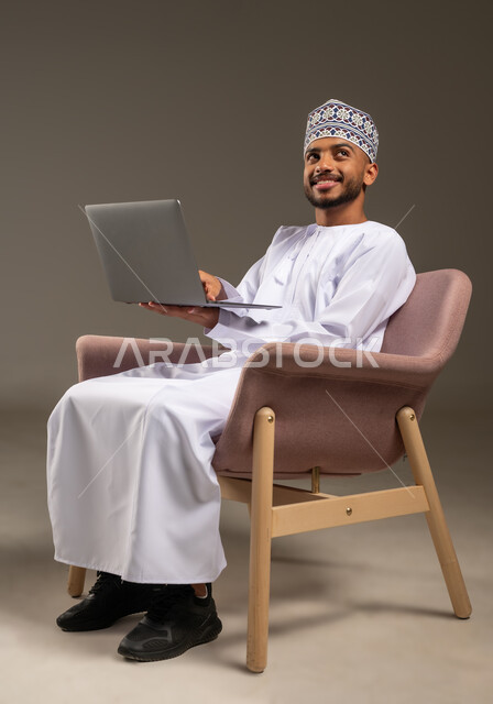 Using advanced modern technologies, managing and organizing work affairs, portrait of an Arab Gulf Omani man wearing a dishdasha and a kummah sitting on a comfortable chair working on a laptop looking up with gestures of pleasure and happiness, browsing social media sites, beige background