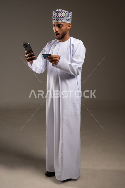 الدفع الإلكتروني عبر التقنيات الحديثة المتطورة، استخدام تطبيقات الجوال للدفع والشراء، بورتريه لشاب عربي خليجي عماني يرتدي الدشداشة والكمة يمسك بيده البطاقة الإلكترونية والهاتف المحمول ينظر بإيماءات الصدمة والمفاجأة, الجسم بالطول الكامل, خلفية بيج