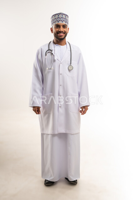 الوقوف باستقامة بإيماءات الثقة بالنفس، العمل في القطاع الصحي في سلطنة عمان، الفحص الطبي وتقديم الخدمات العلاجية للمرضى, بورتريه لطبيب عربي خليجي عماني يرتدي الدشداشة والكمة والمعطف الطبي يضع السماعة الطبية ينظر إلى الكاميرا، الجسم بالطول الكامل، خلفية بيضاء
