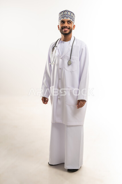 النظر إلى الأعلى بإيماءات الثقة بالنفس، العمل في القطاع الصحي في سلطنة عمان، الفحص الطبي وتقديم الخدمات العلاجية للمرضى, بورتريه لطبيب عربي خليجي عماني يرتدي الدشداشة والكمة والمعطف الطبي يضع السماعة الطبية يقف باستقامة، الجسم بالطول الكامل، خلفية بيضاء
