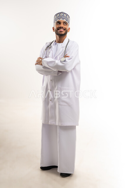 الفحص الطبي وتقديم الخدمات العلاجية للمرضى, بورتريه لطبيب عربي خليجي عماني يرتدي الدشداشة والكمة والمعطف الطبي يضع السماعة الطبية يقف بأيدي متقاطعة ينظر إلى الأعلى بإيماءات الثقة بالنفس، العمل في القطاع الصحي في سلطنة عمان، الجسم بالطول الكامل، خلفية بيضاء