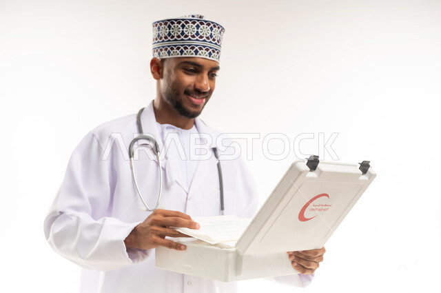 الأدوات والمعدات الطبية اللازمة، مبادئ التعامل مع حالات الطوارئ، بورتريه مقرب لطبيب عربي خليجي عماني يرتدي الدشداشة والكمة والمعطف يحمل حقيبة الاسعافات الأولية المفتوحة، العمل في القطاع الصحي، تقديم الخدمات الصحية الطارئة، خلفية بيضاء