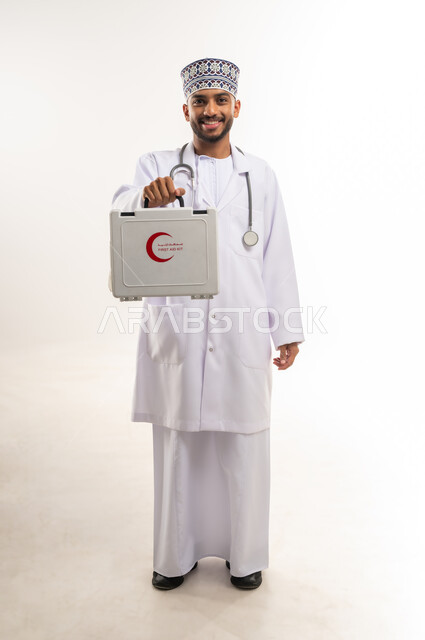 مبادئ التعامل مع حالات الطوارئ، بورتريه لطبيب عربي خليجي عماني يرتدي الدشداشة والكمة والمعطف يحمل حقيبة الاسعافات الأولية ينظر إلى الكاميرا، العمل في القطاع الصحي، تقديم الخدمات الصحية الطارئة، الأدوات والمعدات الطبية اللازمة، الجسم بالطول الكامل، خلفية بيضاء