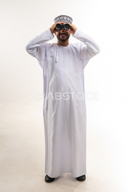 Love of exploration and adventure, portrait of a smiling young Omani Arab Gulf man wearing a dishdasha and a kummah holding binoculars looking at something, gestures of enjoyment and pleasure, seeing distant things through a telescope, full-length body image, white background