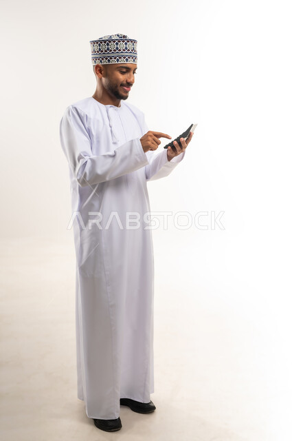 استخدام الحاسبة الرقمية في الأعمال الاقتصادية، محاولة إدارة المصروف المنزلي بتدبر، بورتريه لرجل عربي خليجي عماني يرتدي الدشداشة والكمة يقوم بالحسابات بإيماءات التركيز، السعي نحو التوفير والادخار، الجسم بالطول الكامل، خلفية بيضاء