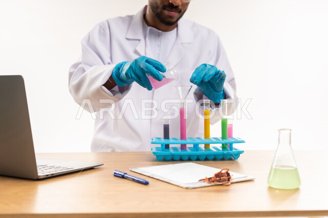 أهمية القيام بتجارب كيميائية، استخدام الحاسوب المحمول في البحث والتطوير، بورتريه مقرب لأخصائي مختبر عربي خليجي عماني يرتدي الزي الخاص بالمهنة يقوم بوضع المحلول داخل الأنبوب المخصص، الاستفادة العملية من البحث المخبري، خلفيه بيضاء