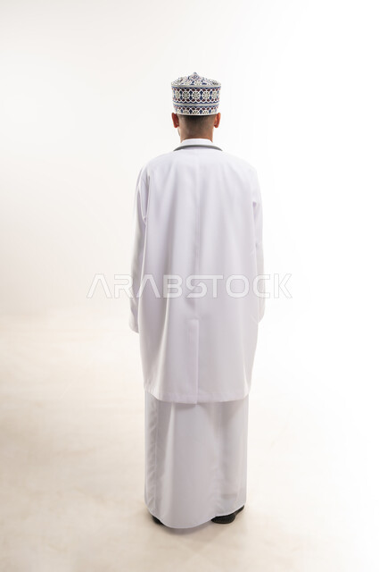 Working in the health sector in the Sultanate of Oman, medical examination and providing treatment services to patients, portrait from the back of an Arab Gulf Omani doctor wearing a dishdasha, a cummerbund and a medical coat standing straight, full body, white background