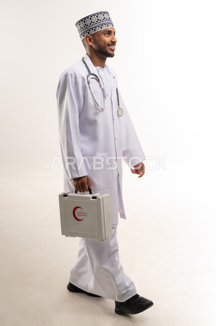 تقديم الخدمات الصحية الطارئة، مبادئ التعامل مع حالات الطوارئ، بورتريه لطبيب عربي خليجي عماني يرتدي الدشداشة والكمة والمعطف يحمل حقيبة الاسعافات الأولية يمشي إلى مكان ما، العمل في القطاع الصحي، الأدوات والمعدات الطبية اللازمة، الجسم بالطول الكامل، خلفية بيضاء