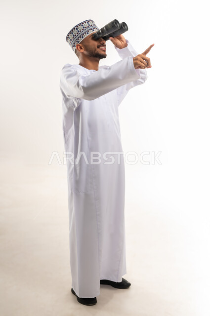 Love of exploration and adventure, portrait of a smiling young Omani Arab Gulf man wearing a dishdasha and a kuma holding binoculars in his hand, pointing his index finger upwards, looking at something, gestures of enjoyment and pleasure, seeing distant things through a telescope, full-length body image, white background