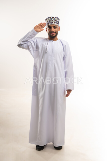 إلقاء التحية والسلام، الأناقة والوسامة والرجولة، مفهوم التقدير والاحترام، بورتريه لشاب عربي خليجي عماني يرتدي الدشداشة والكمة يرفع يده بجانب رأسه ينظر إلى الكاميرا بإيماءات السرور والترحيب، الجسم بالطول الكامل، خلفية بيضاء