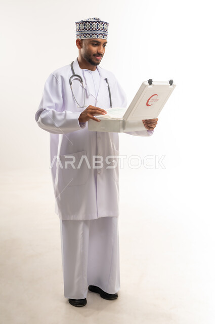 الأدوات والمعدات الطبية اللازمة، تقديم الخدمات الصحية الطارئة للمرضى، مبادئ التعامل مع حالات الطوارئ، بورتريه لطبيب عربي خليجي عماني يرتدي الدشداشة والكمة والمعطف يحمل حقيبة الاسعافات الأولية المفتوحة، العمل في القطاع الصحي، الجسم بالطول الكامل، خلفية بيضاء