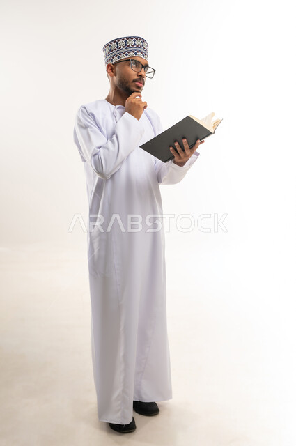 Practicing the hobby of reading and gaining knowledge, interest in browsing useful books, spending free time learning, portrait of a young Arab Gulf Omani man wearing a dishdasha, a turban, and glasses, holding a book in his hand, placing his hand on his chin, looking up with gestures of thinking, full-length body, white background