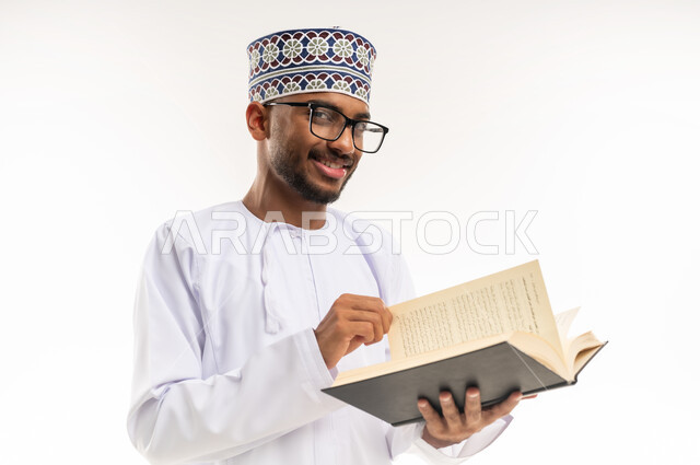 Looking at the camera with gestures of joy and happiness, practicing the hobby of reading and gaining knowledge, spending free time educating and learning, close-up portrait of a young Arab Gulf Omani man wearing a dishdasha, a kimmah and glasses, holding a scientific book in his hand and reading it, white background