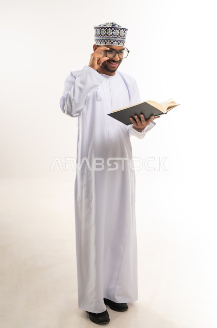 قضاء وقت الفراغ بالتثقيف والتعلم، بورتريه لشاب عربي خليجي عماني يرتدي الدشداشة والكمة والنظارات الطبية يمسك بيده كتاب علمي يقوم بمطالعته بإيماءات التركيز والتمعن، ممارسة هواية القراءة والتزود بالعلم، الجسم بالطول الكامل، خلفية بيضاء