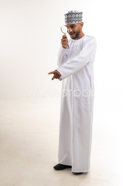 Optical tools for close-up vision, concept of image clarification, pointing with index finger at something, love of exploration and adventure, portrait of a young Arab Gulf Omani man wearing a dishdasha and a kummah holding a magnifying glass in his hand looking through it, viewing small things accurately and clearly, full-length body, white background