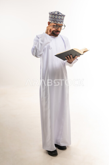 ممارسة هواية القراءة والتزود بالعلم، قضاء وقت الفراغ بالتثقيف والتعلم، بورتريه لشاب عربي خليجي عماني يرتدي الدشداشة والكمة والنظارات الطبية يمسك بيده كتاب علمي يقوم بمطالعته بإيماءات التركيز والتمعن، الجسم بالطول الكامل، خلفية بيضاء