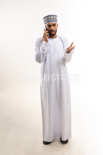 سماع أخبار غير جيدة من الأقارب، الدردشة مع الأهل والأصدقاء، استخدام جهاز تقني حديث، بورتريه لشاب عربي خليجي عماني يرتدي الدشداشة والكمة يقوم بإجراء مكالمة هاتفية عبر الهاتف المحمول يرفع يده بإيماءات الاستياء وعدم الرضا، الجسم بالطول الكامل، خلفية بيضاء