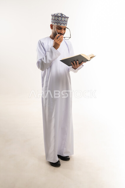Interest in browsing useful books, spending free time learning and educating, portrait of a young Gulf Arab Omani man wearing a dishdasha, a kimono and glasses, holding a book in his hand and reading it with gestures of concentration and contemplation, practicing the hobby of reading and gaining knowledge, full body, white background