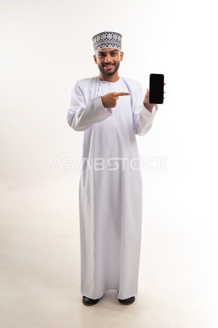 استخدام الأجهزة التقنية الحديثة والمتطورة, النظر إلى الكاميرا بإيماءات السعادة والسرور, بورتريه لرجل عربي خليجي عماني يرتدي الدشداشة والكمة يمسك بيده الهاتف المحمول ويشير إليه بإصبع السبابة, عرض شاشة سوداء فارغة عبر الجوال, الجسم بالطول الكامل, خلفية بيضاء